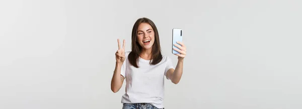 Atrakcyjna Brunetka Wygląda Szczęśliwie Robi Selfie Smartfonie Pokazując Gest Pokoju — Zdjęcie stockowe