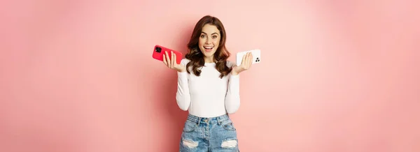 春のプロモーションの概念 スタイリッシュな魅力的な女性は 2つのスマートフォン 両手で携帯電話を保持し 喜んで笑顔 新しい携帯電話 ピンクの背景を購入 — ストック写真