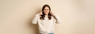 Annoyed and angry woman shut ears, complaining at loud noisy music noise, standing over beige background.