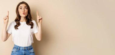 Surprised brunette girl showing promo, pointing and looking up with amazed face expression, standing over beige background.