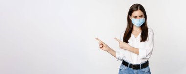 Tıbbi maskeli kurumsal bir kadının portresi, solu işaret ediyor, logoyu, şirket afişini gösteriyor, beyaz arka planda duruyor..