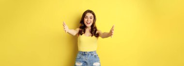 Beautiful girl in tank top, reaching hands, stretching arms forward and smiling, welcome, hugging or greeting someone, standing over yellow background.