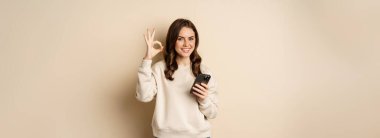 Beautiful woman holding cellphone, mobile phone and okay sign, recommending application, shopping app, standing over beige background.