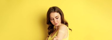 Covid-19 vaccination. Close up portrait of young millennial woman looking at patch on shoulder after vaccine from coronavirus shot, standing against yellow background.