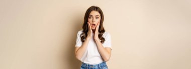 Portrait of stylish young woman gasping, looking surprised and amazed, impressed by smth, standing in t-shirt and jeans over beige background.