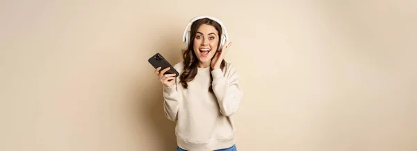 微笑着快乐的女人 在智能手机里听着音乐 兴奋的笑着 站在米色的背景上 — 图库照片