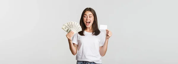 stock image Amused beautiful girl looking at money and holding credit card, standing white background.