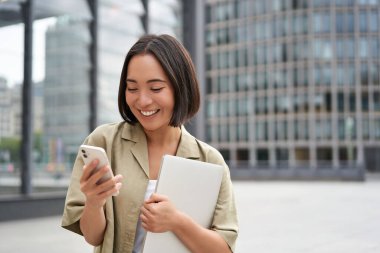 Genç gülümseyen Asyalı kadının portresi sokakta yürüyor, işe dizüstü bilgisayarla ve akıllı telefonla gidiyor..