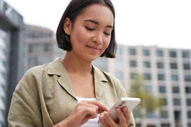 Mobil teknoloji. Gülümseyen Asyalı kadın akıllı telefon uygulaması kullanıyor, caddede telefonuna bakıyor, haritayı kontrol ediyor, birini arıyor ya da mesaj atıyor..