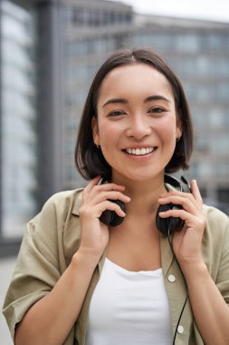 Boynuna kulaklık takmış, gülümseyen ve gülen Asyalı güzel bir kadının gündüz vakti sokakta dikey pozu..