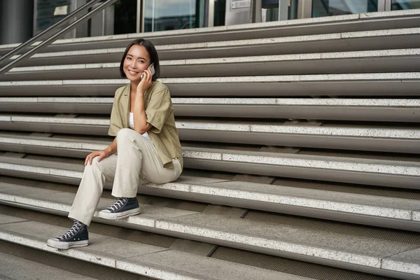 stock image Smiling asian girl sits on stairs of building and talks on mobile phone, relaxing during telephone conversation.