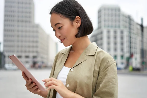 stock image Portrait of asian woman reading, using tablet while standing on street, smiling while looking at screen.