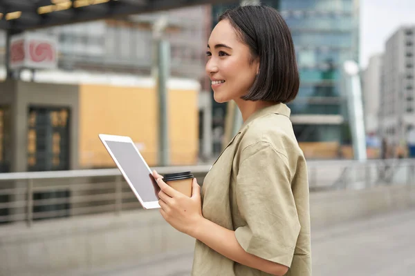 Modern people. Young asian woman with tablet, drinks coffee on street, using app, smiling.