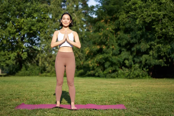 Genç, düşünceli bir kadın, temiz hava üzerine meditasyon yapıyor, parkta yoga egzersizleri yapıyor, spor kıyafetleri içinde lastik minderin üzerinde duruyor..