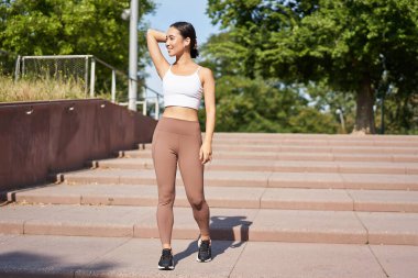 Sporcu kadın dışarıda koşuyor. Kablosuz kulaklıklarla gülümseyen Asyalı fitness kızı müzik dinliyor ve yolda koşuyor..