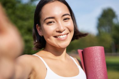 Mutlu Asyalı fitness kızı parkta lastik yoga minderiyle selfie çekiyor. Sağlıklı genç sporcu kadın egzersiz sırasında kendi fotoğrafını çekiyor..