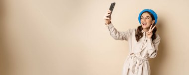 Beautiful modern woman posing for selfie, holding smartphone, taking picture photo on mobile phone, standing happy against beige background.