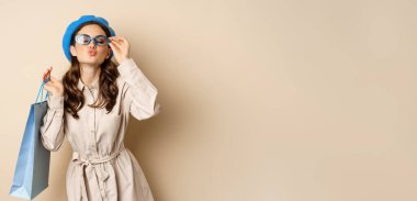 Stylish happy girl on shopping. Portrait of modern woman with shop bag, laughing and smiling satisfied, buying herself gift, standing over beige background.