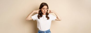 Annoyed young woman shut ears from loud noise, feeling discomfort and complaining, standing against beige background.