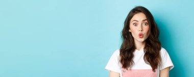 Kıvırcık saç stili ve makyajlı, büzüşmüş dudakları olan, şaşırmış yüzüyle kameraya bakan, mavi arka planda duran heyecanlı stil kızı yakından çek..