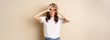 Funny brunette woman having fun, showing finger glasses gesture and fool around, posing over beige background.