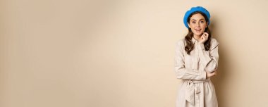 Image of stylsh beautiful woman looking surprised, shocked reaction at camera, posing in trench coat against beige background.