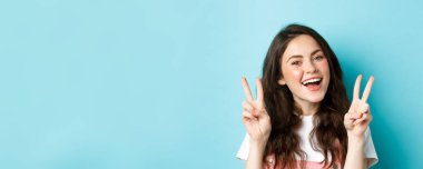 Genç, pozitif, modern bir kız sadece iyi titreşimler yolluyor. V işaretini gösteriyor ve gülümsüyor. Mavi arka planda duruyor..