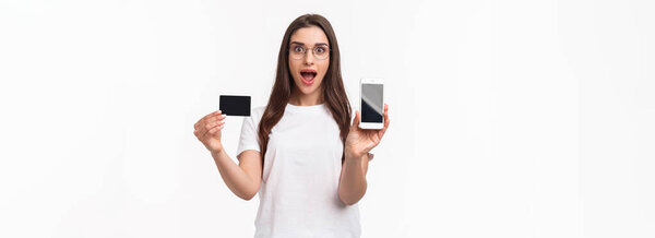 Portrait of excited and happy young woman introduce new application, favorite online store to buy clothes, show mobile phone and credit card, smiling amused, white background.