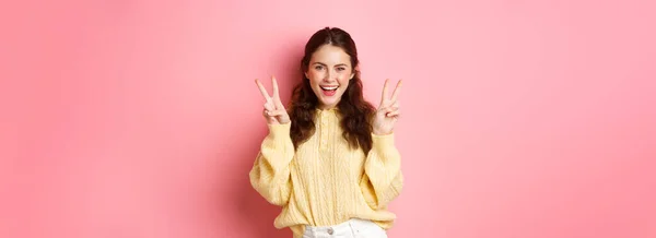 stock image Portrait of attractive brunette female model, smiling and showing peace signs, making v-sign gesture, standing positive and happy against pink background.