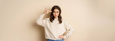 Woman showing finger pistol gesture over her head, being bothered and annoyed, standing over beige background.