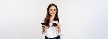 Portrait of young woman looking at smartphone screen, paying for purchase online with credit card, standing over white background, placing order.