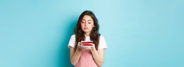 Tatiller ve kutlamalar. Üzgün şirin kız doğum gününü tek başına kutluyor, elinde bir mumla elinde pastayla üzgün üzgün, mavi arka planda tek başına duruyor..