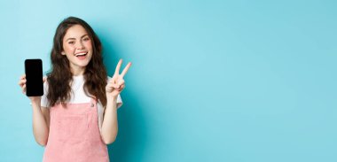 Sevimli genç bir kadın gülümsüyor ve boş akıllı telefon ekranı ile V işareti gösteriyor, uygulamayı veya mobil mağazayı gösteriyor, mavi arka planda duruyor..