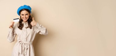 Concept of shopping and sale. Happy young woman in stylish clothes, showing credit card store discount and thumbs up, recommending, standing over beige background.