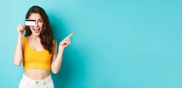 stock image Look there. Stylish modern woman showing plastic credit card, smiling and pointing right, showing way to banner or logo, standing against blue background.