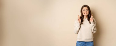 Hopeful worried girl cross fingers, making wish, standing over beige background.