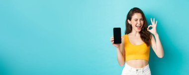 Yaz kıyafetleri içinde çekici bir modern kadın, güven verici bir şekilde göz kırpıyor, boş akıllı telefon ekranıyla işaret dilini gösteriyor, logonuzu ekranda gösteriyor, mavi arka plan..