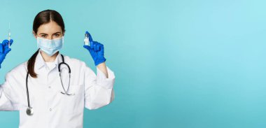 Gülümseyen sağlık personeli, maske takan doktor ve lastik eldiven, mavi arka planda duran covid-19 salgınından şırınga ve aşı gösteriyor..