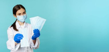 Portrait of medical worker, woman doctor in suggesting face masks from covid-19 pandemic, standing in hospital uniform over torquoise background.