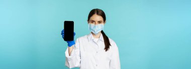 Young woman doctor in medical face mask and hospital uniform, showing mobile phone app, screen interface, online clinic concept, standing over torquoise background.