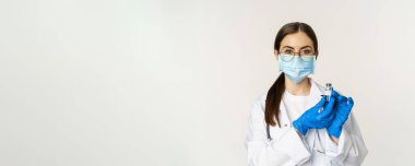 Portrait of young woman, doctor in medical face mask and uniform, showing vaccine, covid-19 vaccination campaign, standing over white background.