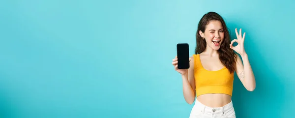 stock image Attractive modern woman in summer clothes, winking assuringly, showing okay sign with blank smartphone screen, showing your logo on display, blue background.