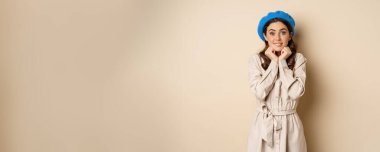 Portrait of beautiful woman in trendy outfit, touching her face and looking dreamy, yearning smth, standing against beige background. People concept