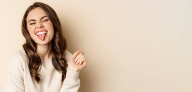 Close up of carefree happy woman, showing tongue and having fun, posing in sweater over beige background. Copy space