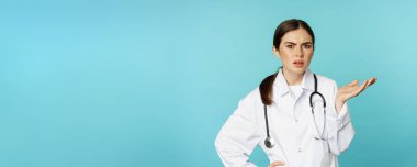 Portrait of annoyed, tired woman doctor, facepalm, roll eyes frustrated, bothered by smth stupid, standing in white coat over torquoise background.