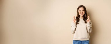 Confident woman praying, making wish with fingers crossed for good luck, standing over beige background.