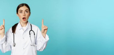 Enthusiastic medical worker, young woman doctor in white coat, stethoscope, showing advertisement, pointing fingers up, standing over torquoise background.