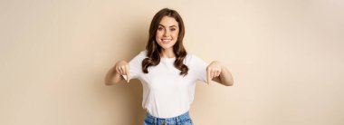 Smiling happy girl pointing fingers down and showing banner, company logo below, click link, standing over beige background.