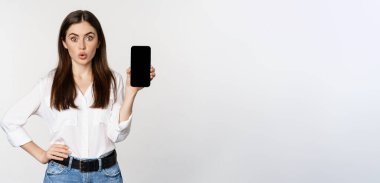 Enthusiastic corporate woman demonstrating website, mobile phone screen, showing application and saying wow, standing over white background.