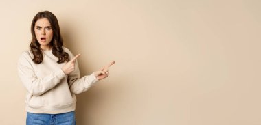 Shocked girl pointing fingers right, gasping startled, standing over beige background.